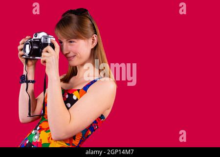 Attraktive junge blonde Frau Tourist mit Sonnenbrille auf dem Kopf lächelt und macht Foto auf Retro-Kamera. Stockfoto