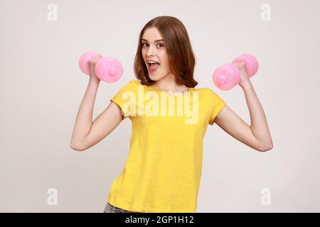 Zufriedene überfröhte Frau im gelben Casual T-Shirt, die mit rosa Hanteln Sportübungen macht, Muskeln aufpumpt und Aufregung ausdrückt. Innenaufnahme des Studios isoliert auf grauem Hintergrund. Stockfoto