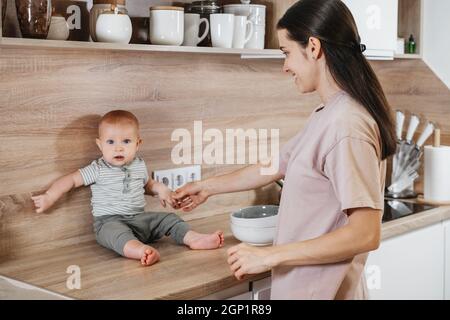 Beschäftigt Mutter Reinigung Küche mit Baby auf den Armen. Frau kocht und hält Säugling, müde Frau macht tägliche Hausaufgaben mit Kind. Stockfoto