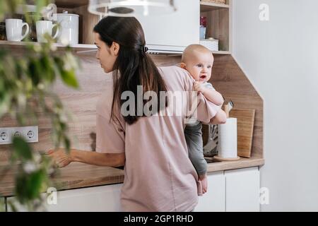 Beschäftigt Mutter Reinigung Küche mit Baby auf den Armen. Frau kocht und hält Säugling, müde Frau macht tägliche Hausaufgaben mit Kind. Stockfoto