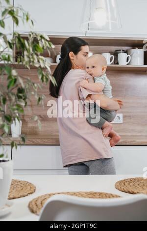 Beschäftigt Mutter Reinigung Küche mit Baby auf den Armen. Frau kocht und hält Säugling, müde Frau macht tägliche Hausaufgaben mit Kind. Stockfoto