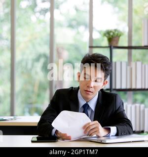 Junger asiatischer Angestellter guckt in den Umschlag. Morgenstimmung im Büro auf einem Tisch mit Tablet und Laptop-Computer. Stockfoto