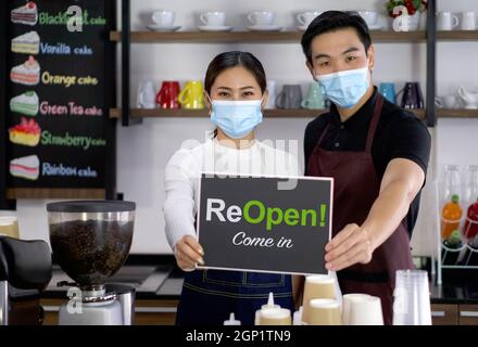 Der Ladenbesitzer und der Barista tragen eine Gesichtsmaske und halten eine Wiedereröffnung! schild vor einem Café-Schalter. Morgendliche Atmosphäre in einem Café. Stockfoto
