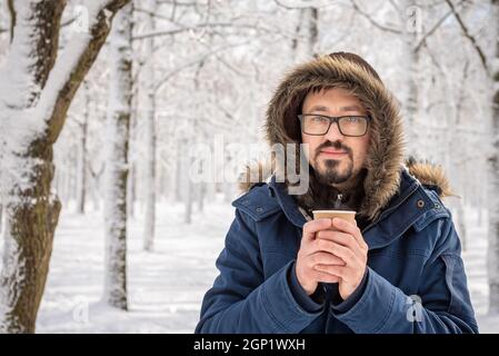 Attraktiver bärtiger Mann in einer Brille, der im Freien in der Kälte des Winters posiert. Straßenportrait im Winter. Kaffee oder Tee in einer Papiertasse in seinen Händen. A man RE Stockfoto