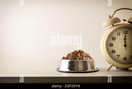 Zeit zum Essen mit einer Schüssel voller trockener Tiernahrung und Uhr daneben auf einem Holztisch und hellem, isoliertem Hintergrund. Vorderansicht. Horizontale Zusammensetzung. Stockfoto