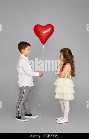 Paar kleine Mädchen und Junge mit roten Herzen Ballons isoliert auf grauem Hintergrund. Junge gibt schüchternes Mädchen Ballon. Valentinstag Stockfoto