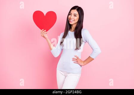 Foto-Porträt des Mädchens zeigt rotes Herz lächelnd in lässigen Kleidern auf pastellrosa Hintergrund isoliert Stockfoto