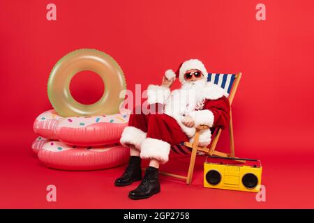 Der weihnachtsmann in Sonnenbrille und Kostüm sitzt auf dem Liegestuhl in der Nähe von Schwimmringen und Boombox auf rotem Hintergrund Stockfoto