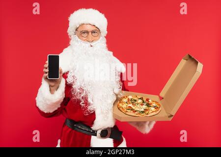 Santa claus zeigt Smartphone mit leerem Bildschirm und hält Pizza in Box isoliert auf rot Stockfoto