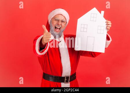 Aufgeregt älterer Mann mit grauem Bart trägt weihnachtsmann Kostüm mit Papierhaus in den Händen zeigt Daumen nach oben, empfehlen Immobilienagentur. Innenaufnahme des Studios isoliert auf rotem Hintergrund. Stockfoto