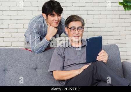 Asiatische Senior Geschäftsmann mit digitalen Tablet diskutieren zusammen mit jungen Team im Büro. Vater Mann und sein Sohn sitzen auf dem Sofa und unterhalten sich im Video Stockfoto