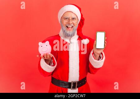 Lächelnder älterer Mann mit grauem Bart im weihnachtsmann Kostüm stehend hält Smartphone mit weißem leeren Bildschirm und Sparschwein, Cashback, Online-Brieftasche. Innenaufnahme des Studios isoliert auf rotem Hintergrund. Stockfoto