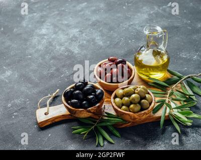 Set aus grünen, schwarzen und roten oder rosa Oliven und Olivenöl auf grauem Hintergrund. Verschiedene Olivenarten in Olivenholzschüsseln und Olivenöl über Holz Stockfoto