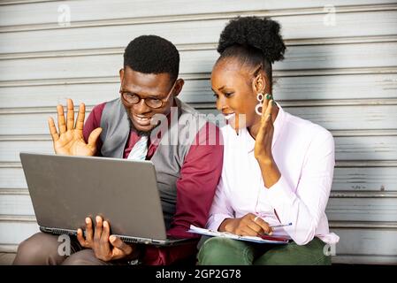 Glückliches junges afrikanisches Geschäftspaar, das draußen sitzt und einen Videoanruf mit einem tragbaren Laptop-Computer hat, der die Hand winkt und lächelt Stockfoto