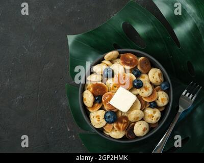 Trendige Küche: Pfannkuchen, Müsli, Butter und Heidelbeeren. Haufen von Mini-Cerealien-Pfannkuchen in Boul auf tropischem Monsterra-Blatt auf dunklem Hintergrund. Cop Stockfoto
