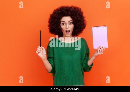 Überrascht Frau mit Afro-Frisur trägt grünen lässigen Stil Pullover stehen mit offenem Mund, hält Papier Notizbuch und Bleistift. Innenaufnahme des Studios isoliert auf orangefarbenem Hintergrund. Stockfoto