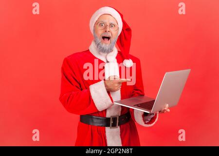 Aufgeregt älterer Mann mit grauem Bart trägt weihnachtsmann Kostüm stehen mit Laptop in den Händen, zeigt auf Computer-Bildschirm mit überrascht aussehen. Innenaufnahme des Studios isoliert auf rotem Hintergrund. Stockfoto