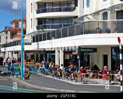 Gäste sitzen in Straßencafés entlang der Marine Parade und genießen die Sonne im August in Mount Maunganui, Bay of Plenty, North Island, Neuseeland. Die entspannte Stadt am Meer ist bekannt für ihren erloschenen Vulkan, den Pazifik-Strand und Straßencafés. Stockfoto