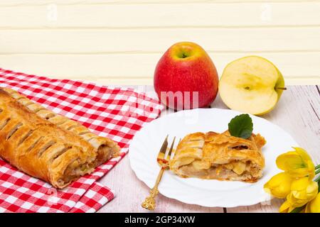Apfelrolle serviert. Eine Rolle hausgemachter frisch gebackener Apfelstrudel mit einer appetitlichen Scheibe Apfelkuchen auf dem Teller, frische Äpfel und Tulpen auf rotem Nickerchen Stockfoto