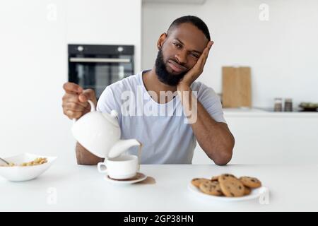 Burnout-Syndrom. Porträt eines erschöpften, verschlaften, tausendjährigen Mannes, der eine weiße Teekane hält, die heißes Getränk aus der Tasse gießt, die heißes Getränk auf der Speisekarte verschüttet Stockfoto