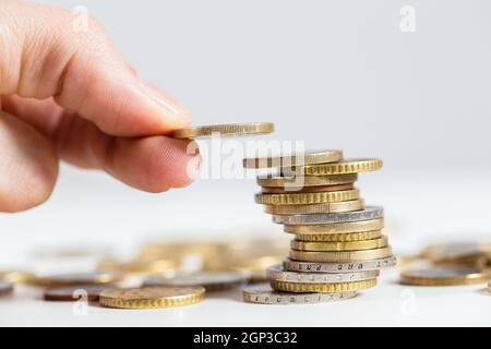 Münze aus der Nähe über den Geldstapel halten. Um einen Euro auf den Haufen metallischen Bargeldes zu erhöhen. Konzept der Einsparung von Gehalt für einen besseren Gewinn. Stockfoto