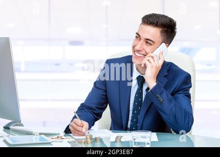 Nahaufnahme eines attraktiven jungen Buchhalters, der im Büro auf dem Smartphone spricht. Junger Mann an der Rezeption vor dem Computer, der Finanzunterlagen ausfüllte Stockfoto
