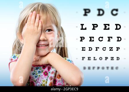 Niedliche kleine Mädchen, die Überprüfung der Sehkraft. Mädchen schließen ein Auge mit der Hand lesen Druckbuchstaben auf Vision Diagramm mit Fokus-Punkt. Stockfoto