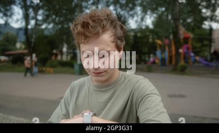 Der junge, blonde, mit bunten Augen beäugte Junge schaut auf seine Armbanduhr und vermittelt, dass die Zeit zu spät ist, schaut auf die Kamera und schüttelt den Kopf Stockfoto