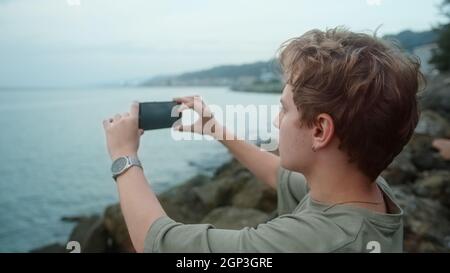 Junger blonder Junge, der den Sonnenuntergang und das Meer über der Bucht von oben auf dem Stein beobachtet, macht Panoramafotos oder nimmt Videos am Telefon auf Stockfoto