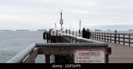 Ein Paar geht auf dem Seal Beach Pier, während Containerschiffe im Hintergrund in Seal Beach, Kalifornien, USA, anstehen Stockfoto