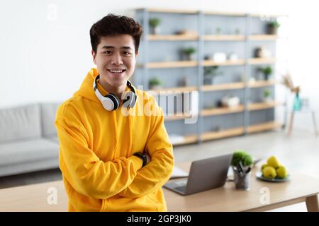 Erfolgreiche Person. Porträt eines selbstbewussten lächelnden asiatischen Mannes in knallgelbem Hoodie und Headset, der auf dem Schreibtisch sitzt, mit gefalteten Armen posiert und lo Stockfoto