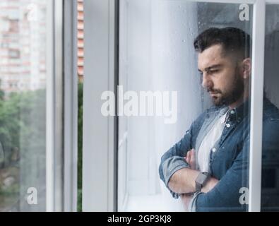 Depressiver kaukasischer Mann verliert Job und ist untröstlich, allein in der Nähe des Fensters. PTBS, psychische Gesundheit und Depressionen. Traurige unglückliche junge gutaussehende Kerl mit gekreuzt Stockfoto