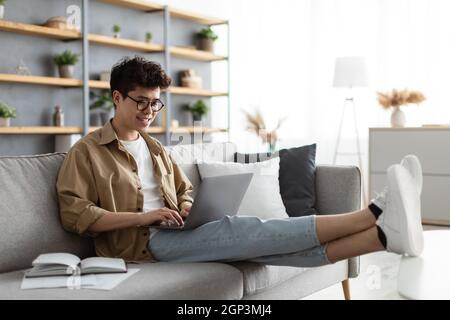 Junger, zwangloser asiatischer Kerl mit Laptop, der im Wohnzimmer auf dem Sofa sitzt, freier Platz zum Kopieren. Glücklicher Mann in einer Brille, der online mit Freunden chattet, männliche Freela Stockfoto