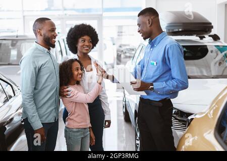 Nettes afro Mädchen hoch fiving positive Verkäufer bei Autohaus. Freundliche schwarze Familie Kauf oder Vermietung eines neuen Autos, die Vereinbarung mit dem Verkäufer bei einem Stockfoto