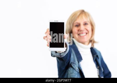 Reife Frau zeigt ein Smartphone, das es in der Hand hält. Weißer Hintergrund. Der Telefonbildschirm ist leer. Bereit für Mockup oder Vorlage. Stockfoto
