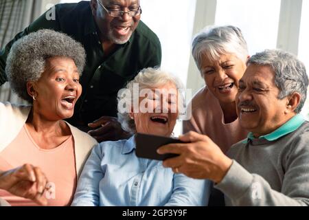 Fünf glückliche, vielfältige ältere Freunde sitzen auf dem Sofa und schauen sich das Smartphone an Stockfoto