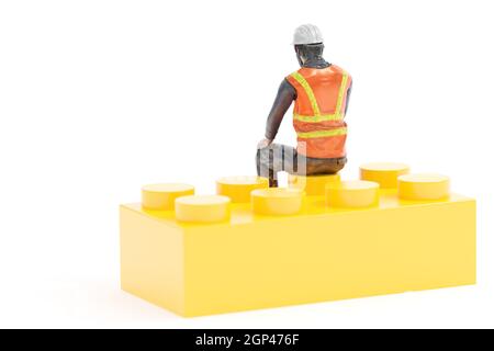 Miniaturarbeiter mit Sicherheitsausrüstung auf gelbem Spielzeugstein vor weißem Hintergrund Stockfoto