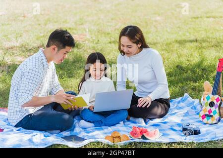 Happy Asian Lifestyle junge Familie Vater, Mutter und kleines Mädchen mit Spaß im Freien sitzen auf Picknickdecke mit Laptop-Computer-Technologien while Stockfoto