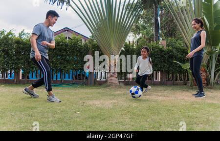 Asiatische junge Mutter, Vater und Kind Tochter Fußball spielen draußen in der Natur ein Feldgarten Park. Glückliche Familie Kind lustig spielen Fußball zusammen in s Stockfoto