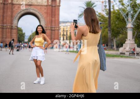 Junge weibliche Reisende in stilvollem Kleid, die Fotos von lächelnden asiatischen Freundinnen auf dem Handy machen, während sie während der Sommerferien die Sehenswürdigkeiten der Stadt Barcelona erkunden Stockfoto