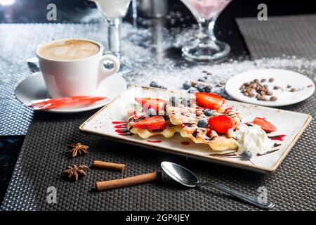 Französisches Dessert – knusprige Waffeln mit Eis, Frischkäse und Marmelade. Obststücke zum Nachtisch. Cappuccino in einem weißen Becher. Stockfoto