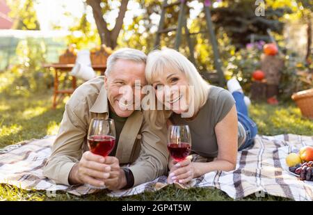 Porträt eines liebenden älteren Paares, das draußen Wein trinkt, auf einer Decke liegt, während er im Garten picknickt und die Kamera anlächelt. Glückliche ältere Ehepartner ce Stockfoto