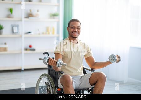 Körperliche Aktivitäten für querschnittsgelähmende Menschen. Beeinträchtigter schwarzer Mann im Rollstuhl, der zu Hause Reha-Übungen mit Hanteln macht. Behinderter junger Mann, der trainiert Stockfoto