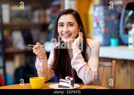 Köstliches Dessert. Glückliche arabische Dame, die Kuchen aß und Kaffee trank, während sie sich im Café ausruhte und die Kamera anlächelte, freien Platz. Junge Frau genießt süß Stockfoto