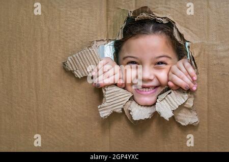 Lächelndes Mädchen hinter einer Pappe, das durch ein großes Loch in der Pappe und mit den Händen am Rand des Lochs schaut Stockfoto