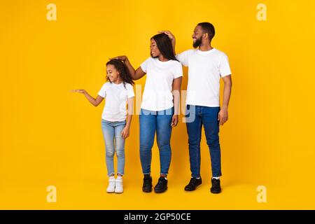 Foto in voller Körperlänge von drei Familienmitgliedern, die die Höhe mit der Handfläche messen. Lächelnde Mutter, Vater und Tochter, die sich bei der Entwicklung anfeuern Stockfoto