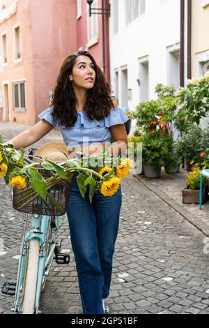 Schöne junge Dame, die durch die Altstadt läuft, während sie ihr türkisfarbenes Fahrrad mit Sonnenblumen in einem Korb schiebt. Sonnenstrahl im Hintergrund. Stockfoto
