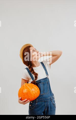 Die Bäuerin in Overalls hält einen großen Kürbis und schaut mit einem Weidenhut nach oben. Stockfoto