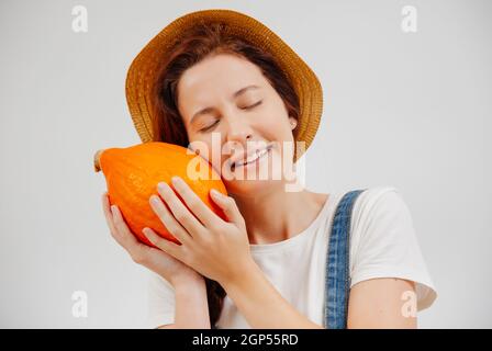 Frau in Overalls und einem Hut hält einen reifen orangefarbenen Kürbis fest. Stockfoto