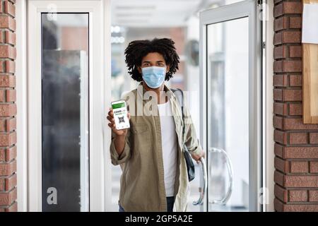 Schwarzer Mann In Schützender Medizinischer Maske Zeigt Digitales Impfzertifikat Beim Betreten Des Gebäudes, Junger Afroamerikanischer Mann Demonstriert Smartph Stockfoto